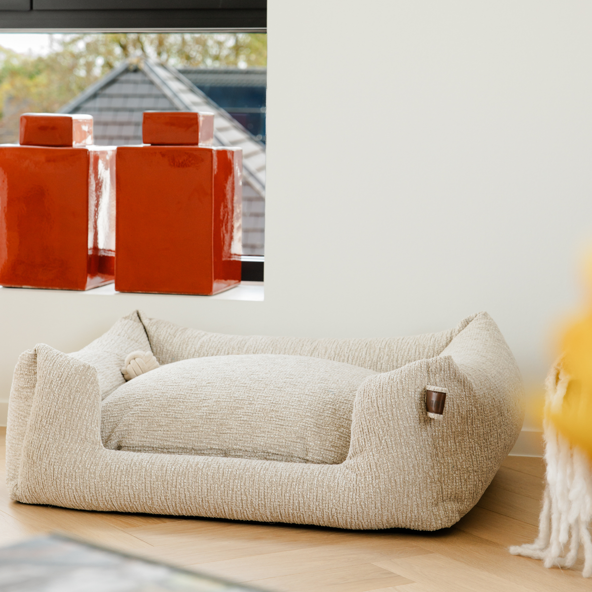 A textured cream-colored dog bed with a raised border and plush cushion, set on a light wood floor against a white wall..#color_clay-beige