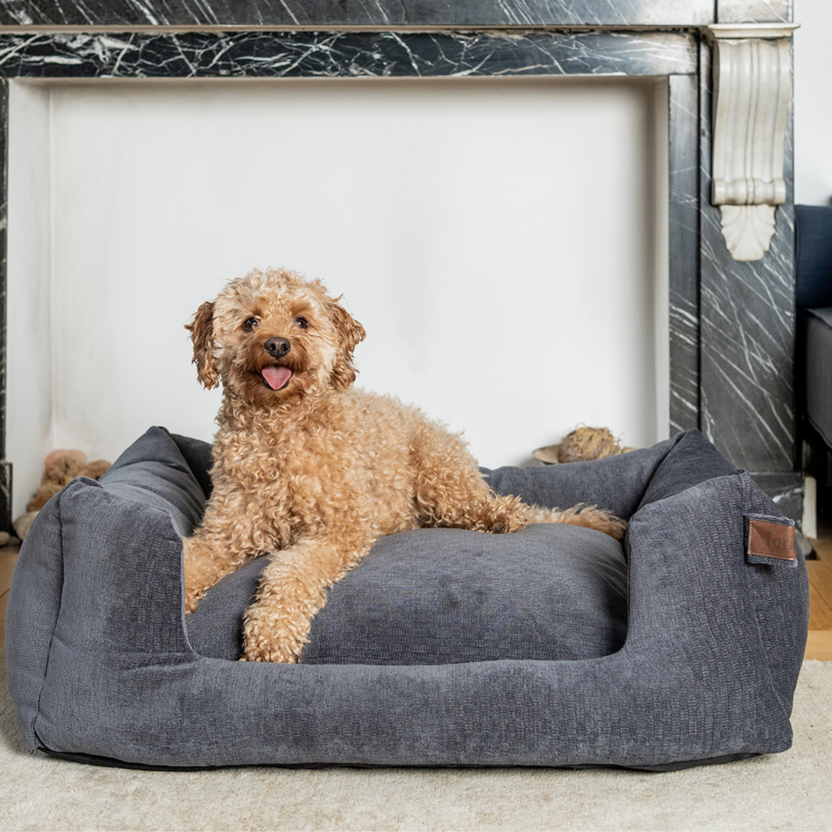 A fluffy light brown dog lies on a large dark gray fabric pet bed placed in front of a white fireplace opening with a marble mantel..
