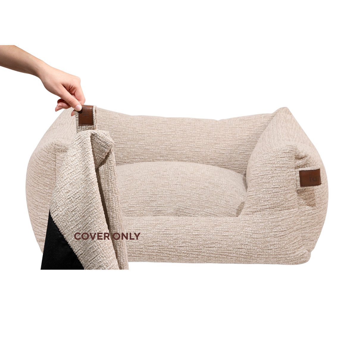 A beige textured pet bed with brown leather-like corner accents, shown on a white background with a hand lifting the cover to reveal a black interior.#color_clay-beige