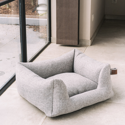 A light gray bouclé pet bed with a raised, cushioned design, placed on a light beige tiled floor next to a large glass window.