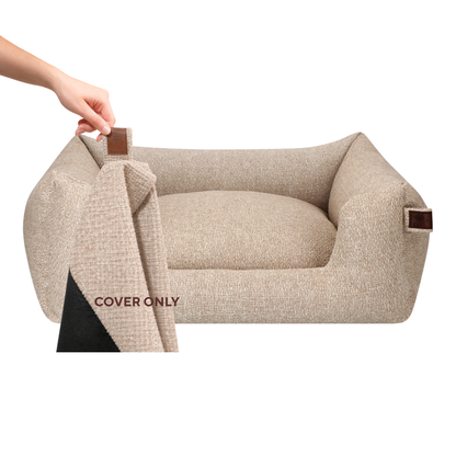 Beige textured pet bed cover shown on a raised pet bed, with a hand lifting the corner to reveal the black underside. A close-up inset at the bottom left shows the fabric texture and a brown tab detail.