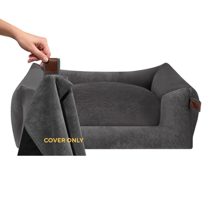 A dark gray pet bed with a matching fabric cover draped off one side, held by a person's hand. The cover has a small brown leather tab, and the bed features small brown leather accents on its sides.