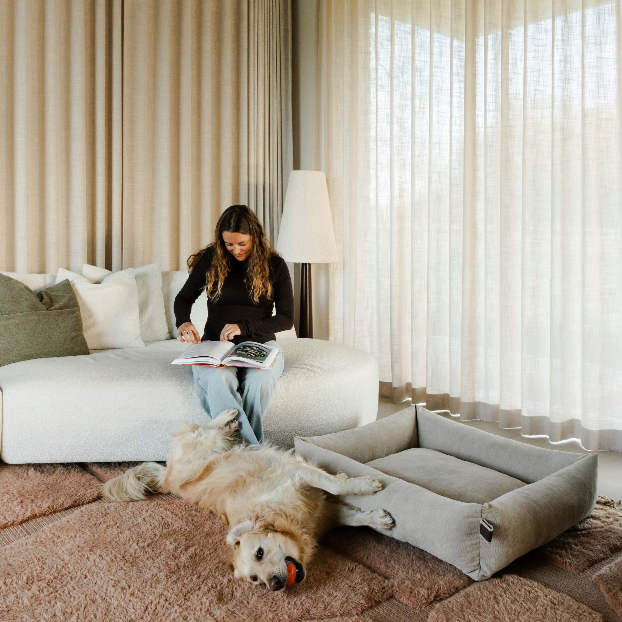 A dog asleep in the Oat Beige Snug dog bed while its owner reads nearby 